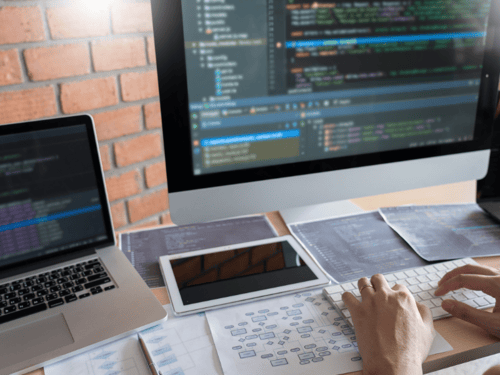 Developer working on code with multiple devices, including an iMac, MacBook, and a tablet, displaying an iOS development environment.
