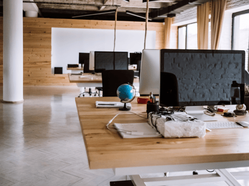 Modern office space with workstations equipped with computers and supplies, illuminated by large windows.