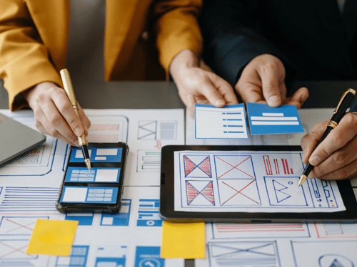 Two designers working on mobile and tablet interface layouts using wireframes and mockups on paper and digital devices.