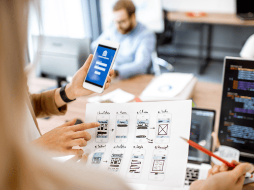 Mobile app development timeline: team reviewing UI wireframes and testing a prototype on a smartphone in a modern office setting.